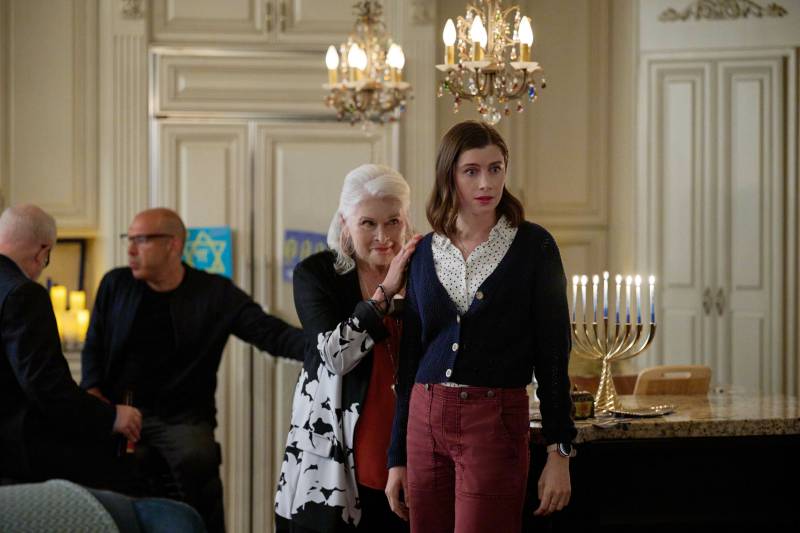 A young woman casually dressed stands in a dining room with two small chandeliers hanging above. Behind her is an elderly woman pushing on her shoulder. Jewish symbols of faith are visible in the background.