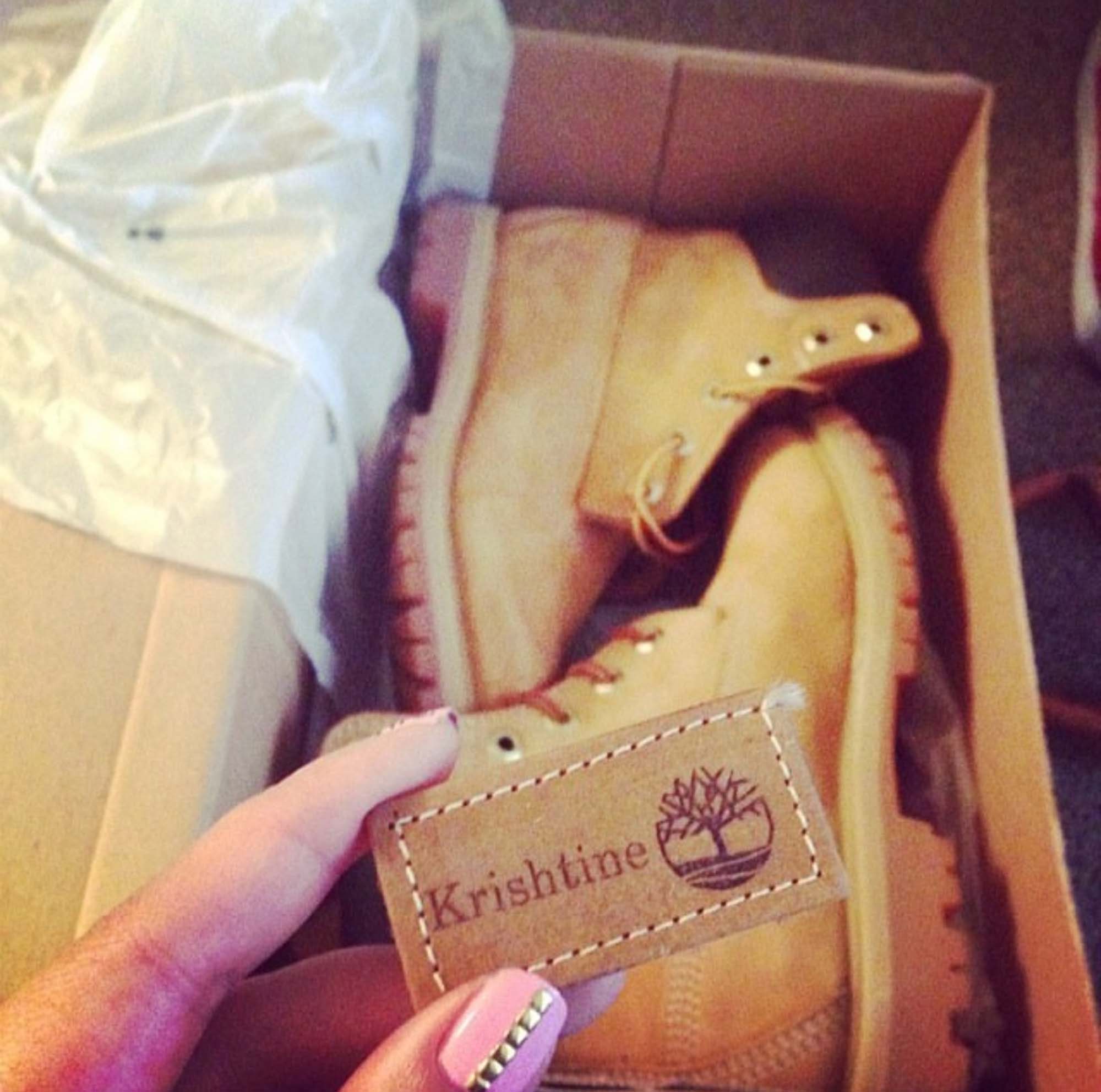 Over a box a Timberland boots, a hand holds up an engraved leather name tag that reads "Krishtine"