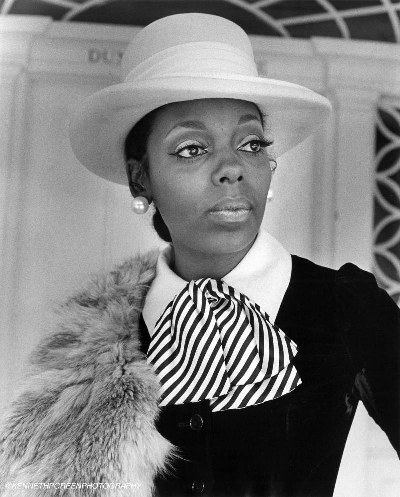 Black and white photo of a Black women in a white hat and stylish clothes draping a fur over her shoulder