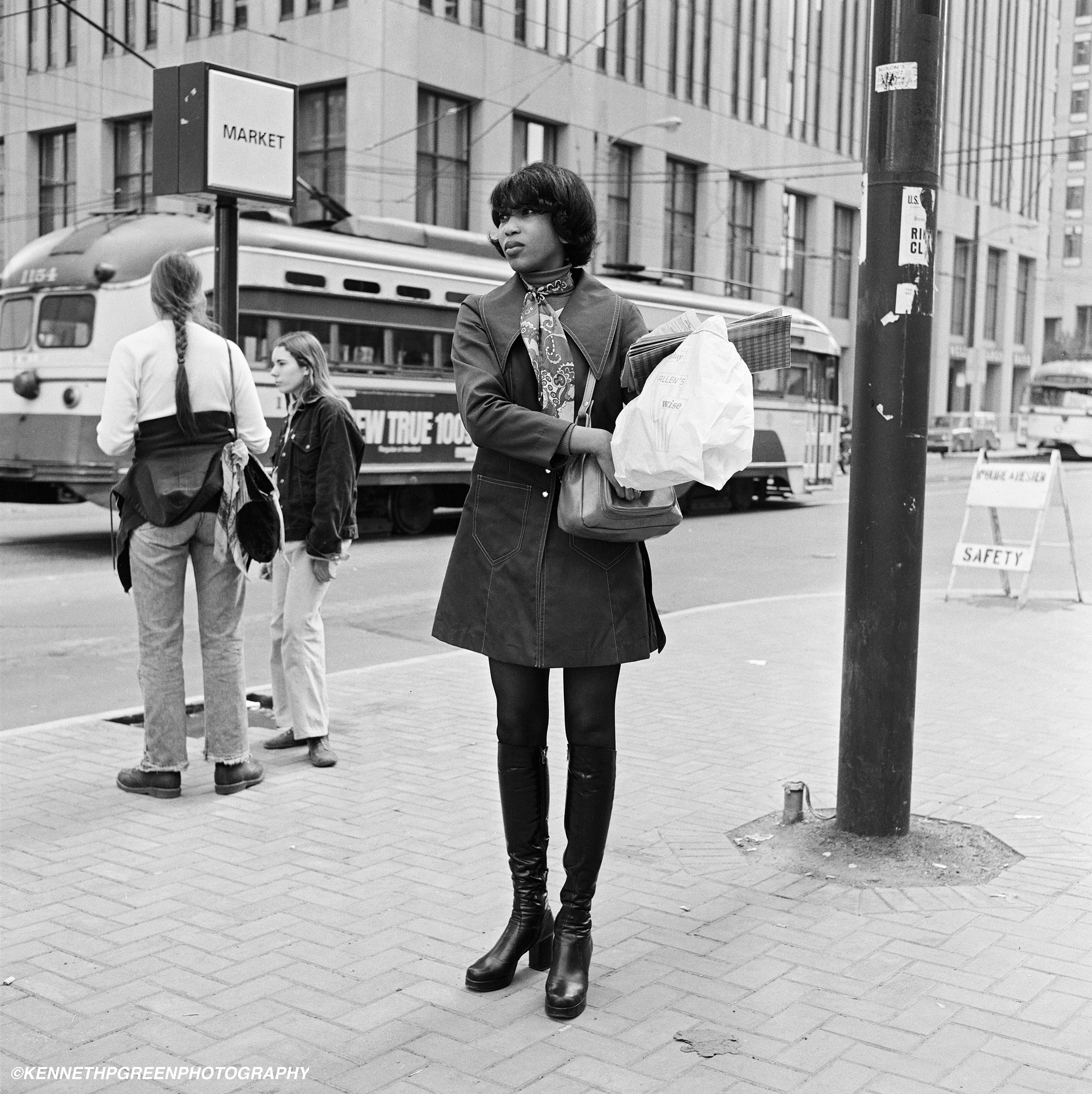 A woman wearing black boots and holding a bag stand on the sidewalk surrounded by buildings.