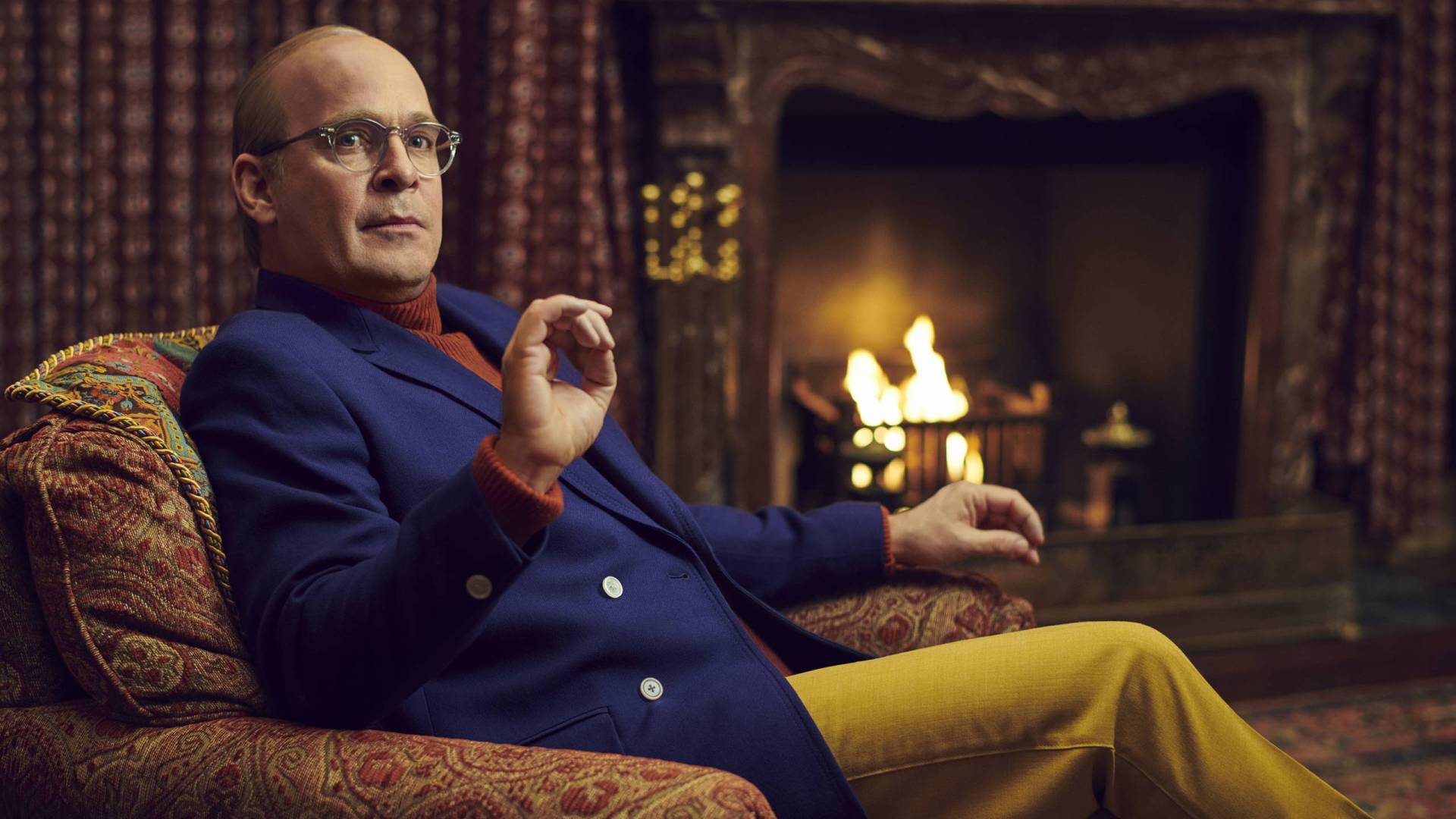 A refined man in a colorful 1970s-era suit sits before a fireplace. He is bald and wearing glasses.