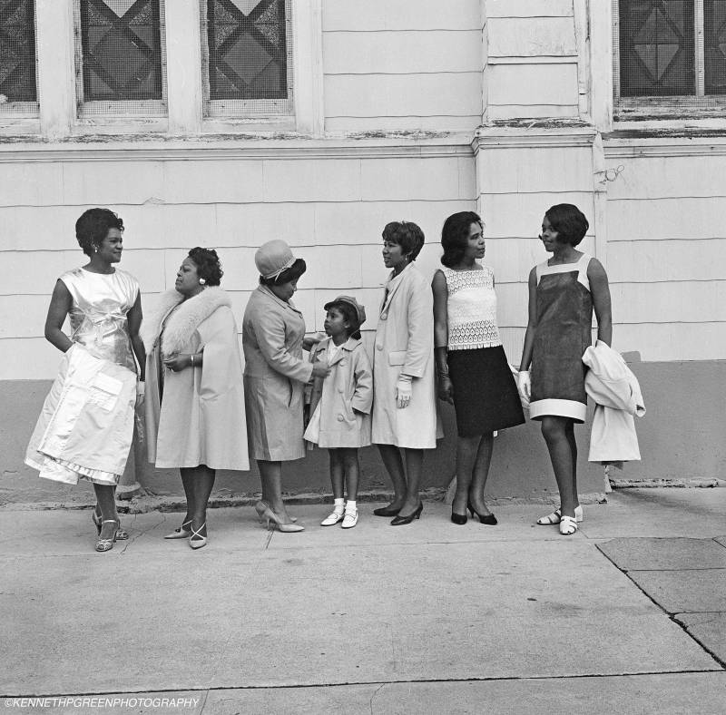 Seven nicely dressed Black women of various ages stand in a row on the street outside a building.