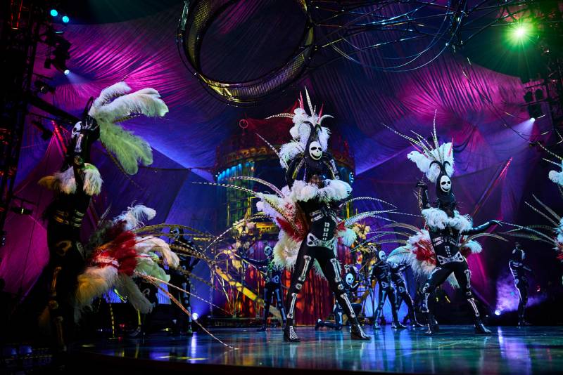 Performers in skeleton costumes with feather headdresses stand on stage.