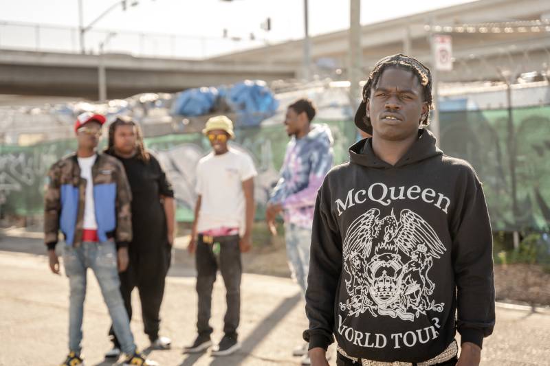 A rapper poses in front of a graffitied fence, with a group of friends standing in the background behind him.