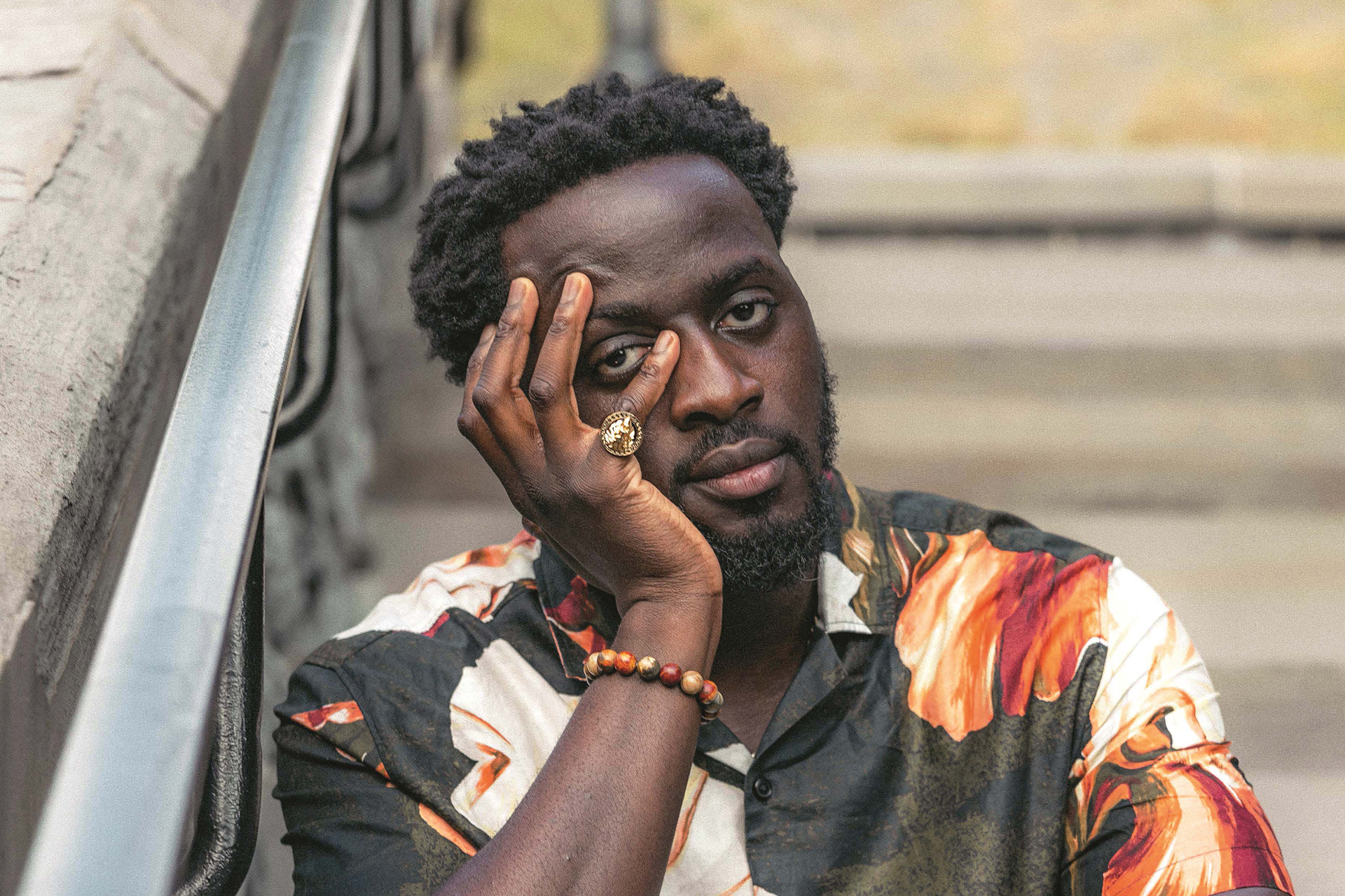 Nana Kwame Adjei-Brenyah sits on a staircase while looking at the camera with one hand on his face.