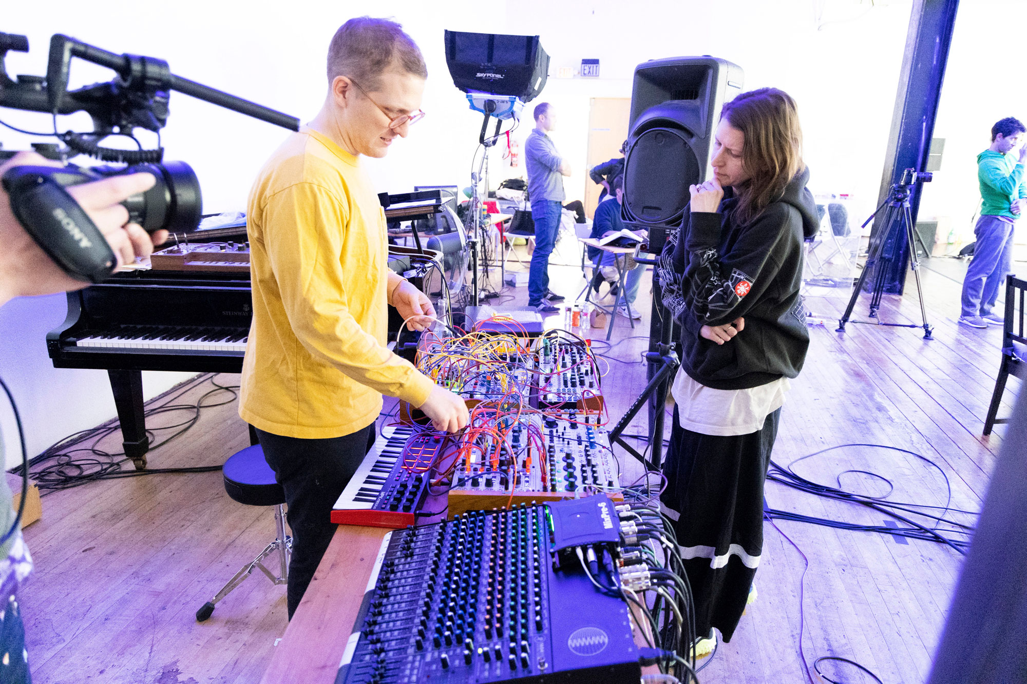 Person in yellow shirt and person in black hoodie stand over synthesizer covered in many colored cables