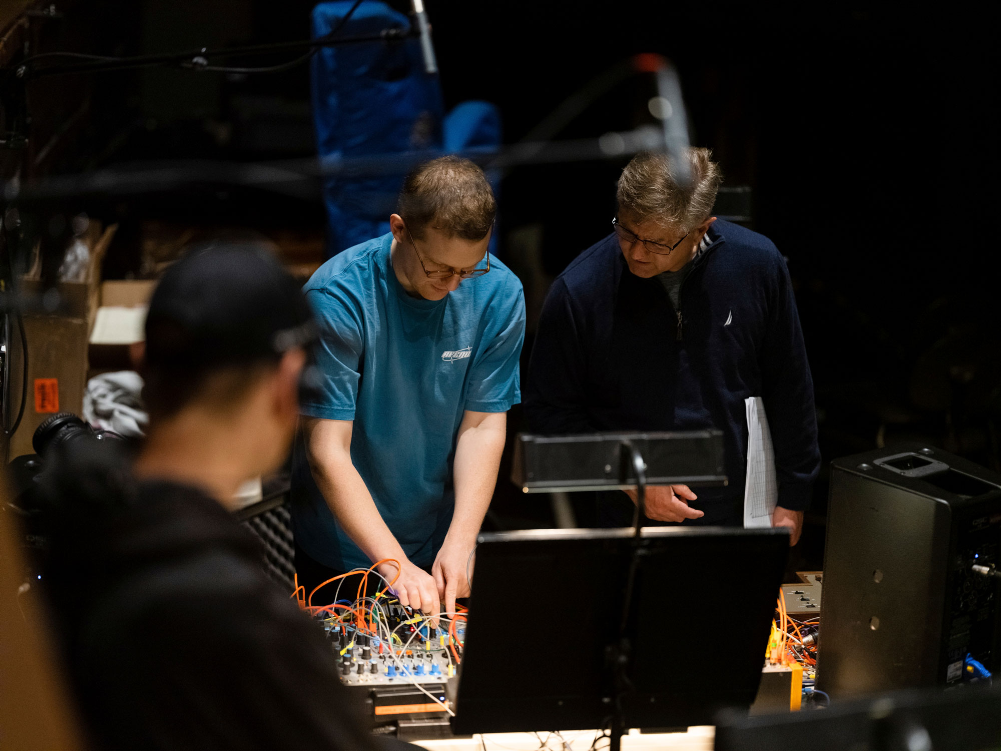 Two men lean over synthesizer with many colored cables