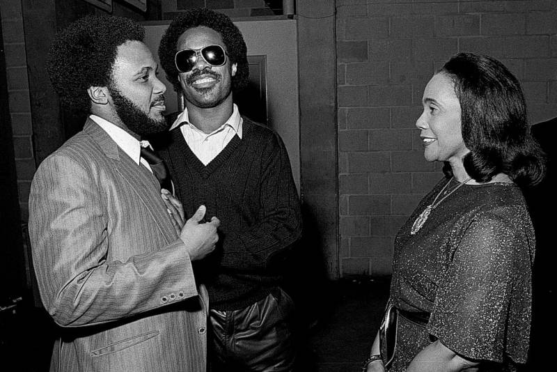 A black-and-white photo of Stevie Wonder smiling and conversing with Coretta Scott King and Martin Luther King III.