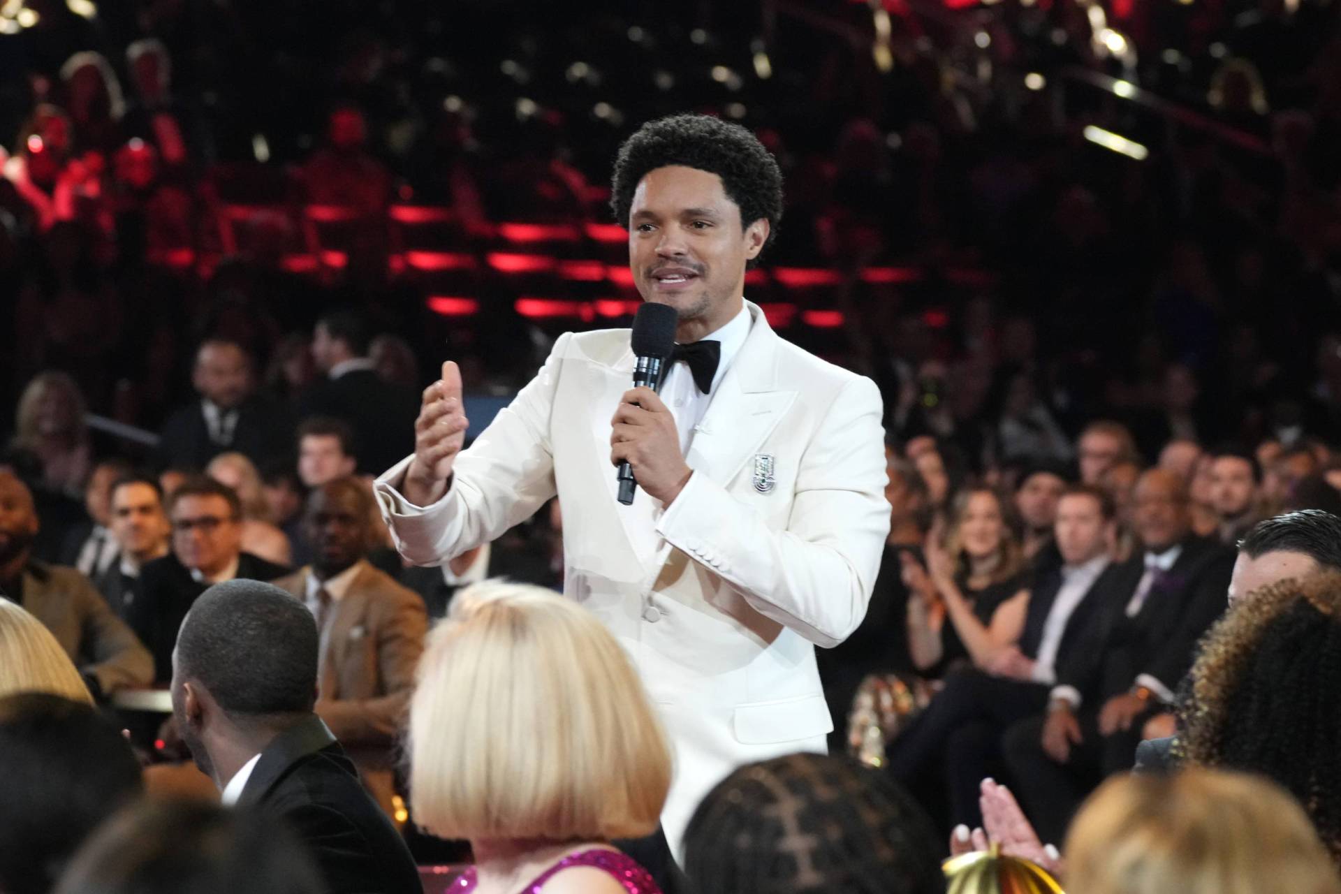 A Black man in a white tuxedo stands among tables full of people, speaking into a microphone.