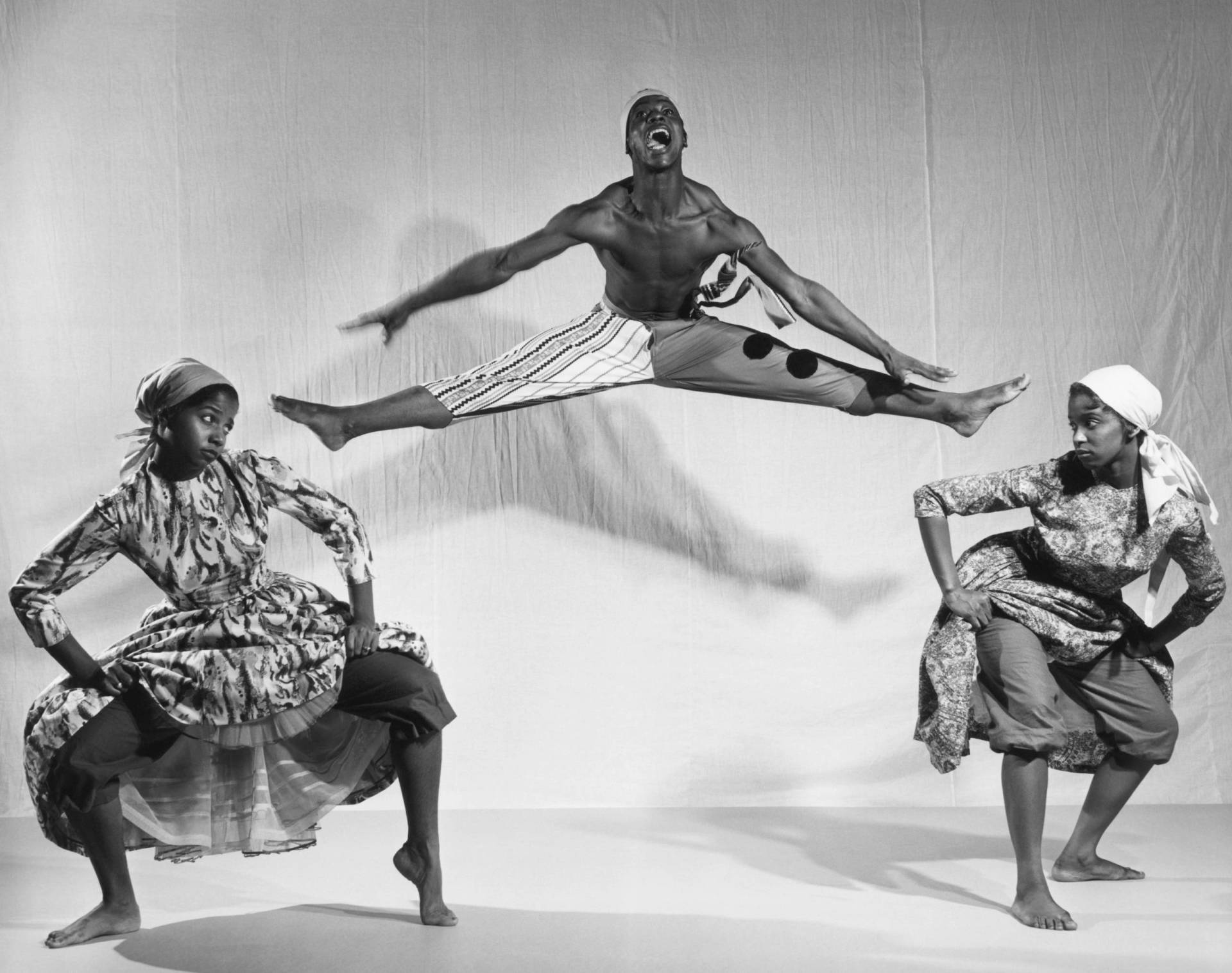 A Black male dancer does the splits in mid-air, while two Black women dance either side of him.