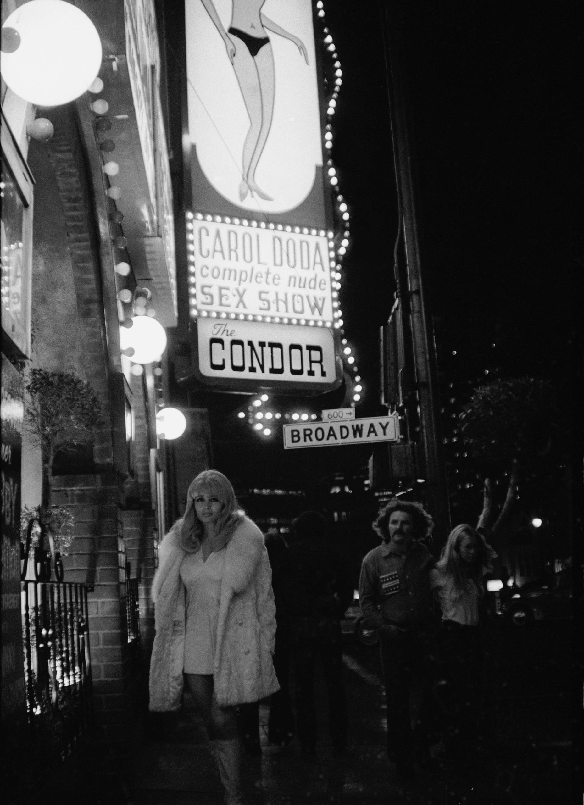 A beautiful blond woman wearing a white coat, miniskirt and boots under a marquee advertising Carol Doda at the Condor.