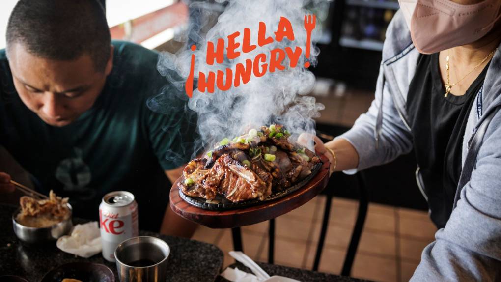 A masked server brings an order of galbi (Korean grilled short ribs) to the table when a man hunches over his food. A logo in the middle of the image reads, "¡Hella Hungry!"