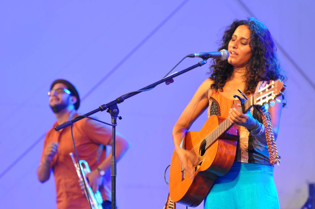 a woman of Indian descent with dark curly hair and a turquoise dress plays guitar on stage in front of a purple background, while a man holds a trumpet near her
