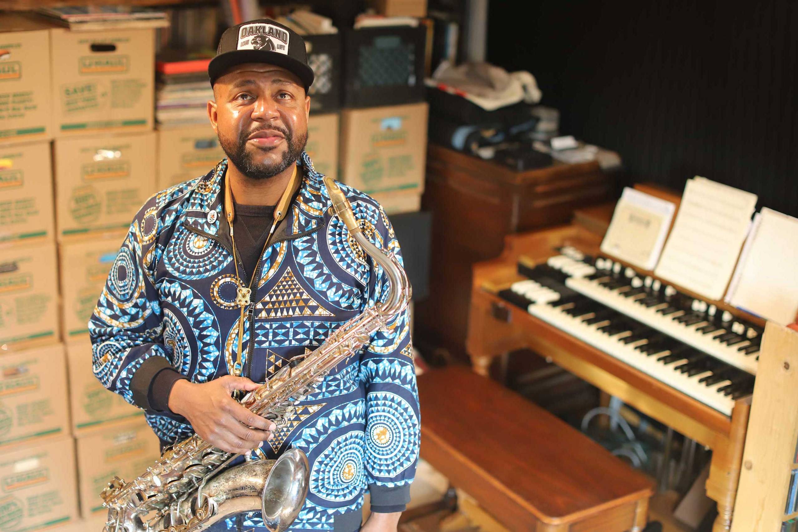 A Black man in a cap and patterned blue shirt stands with a saxophone, with moving boxes and an organ in the background