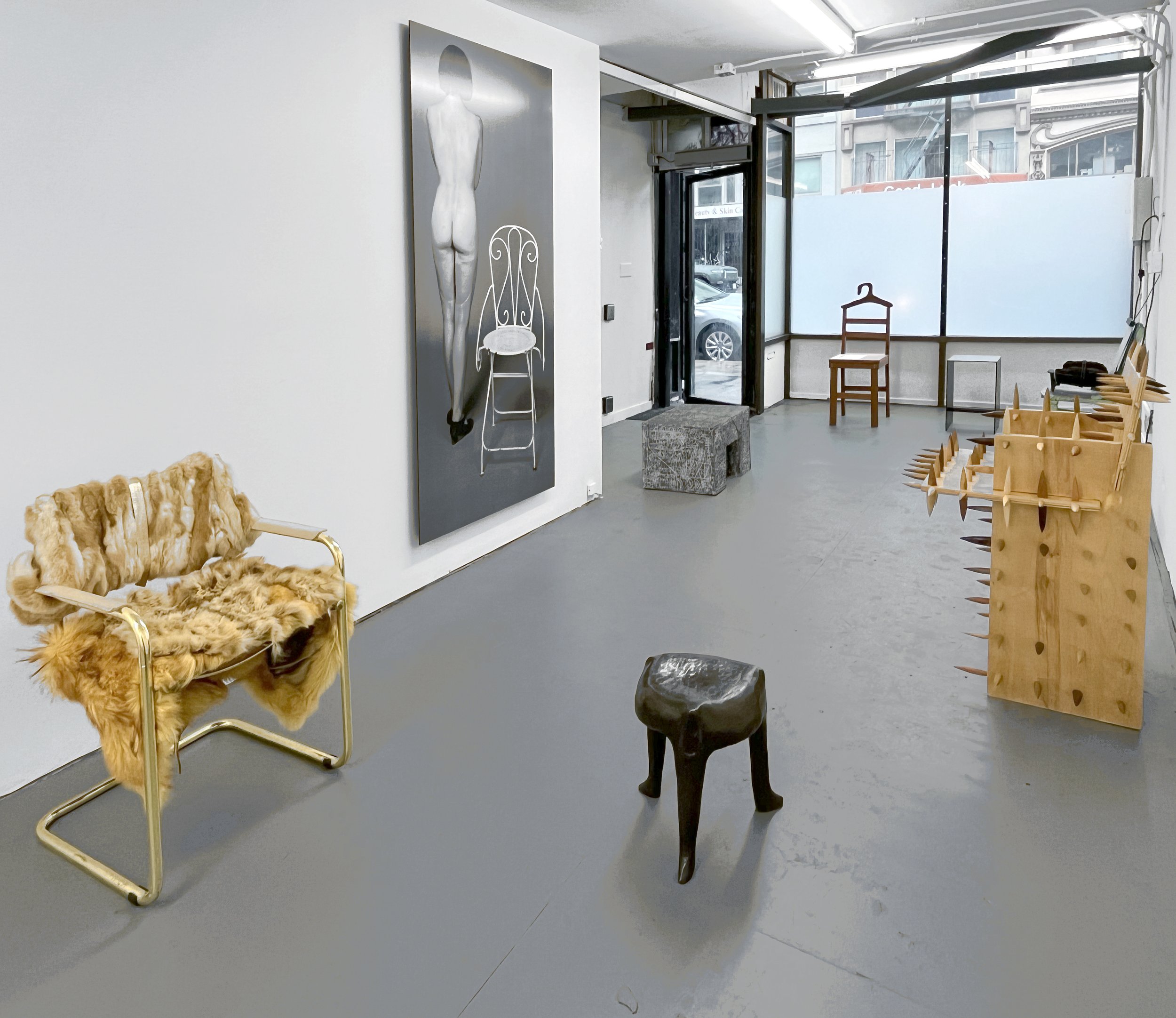 Various chairs and large photograph of nude figure nexts to chair in dark-gray floor white wall gallery space