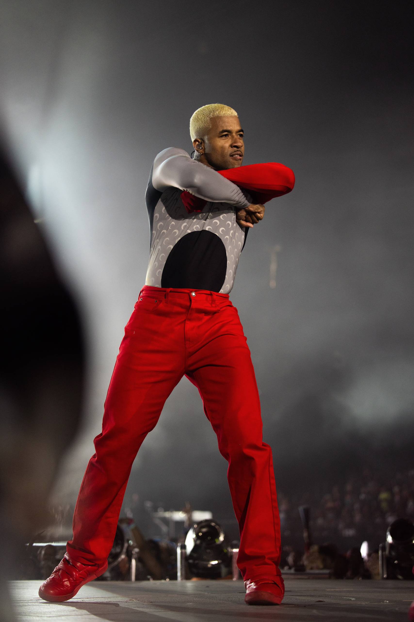 A dancer with bleached hair folds his arms o0n stage while wearing a silver top and red pants.