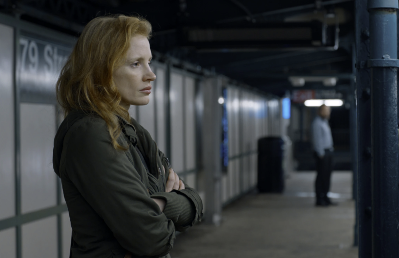 A pale woman with long red hair stands on a subway platform, her arms folded in front of her. A man can be seen in the distance, looking over at her.