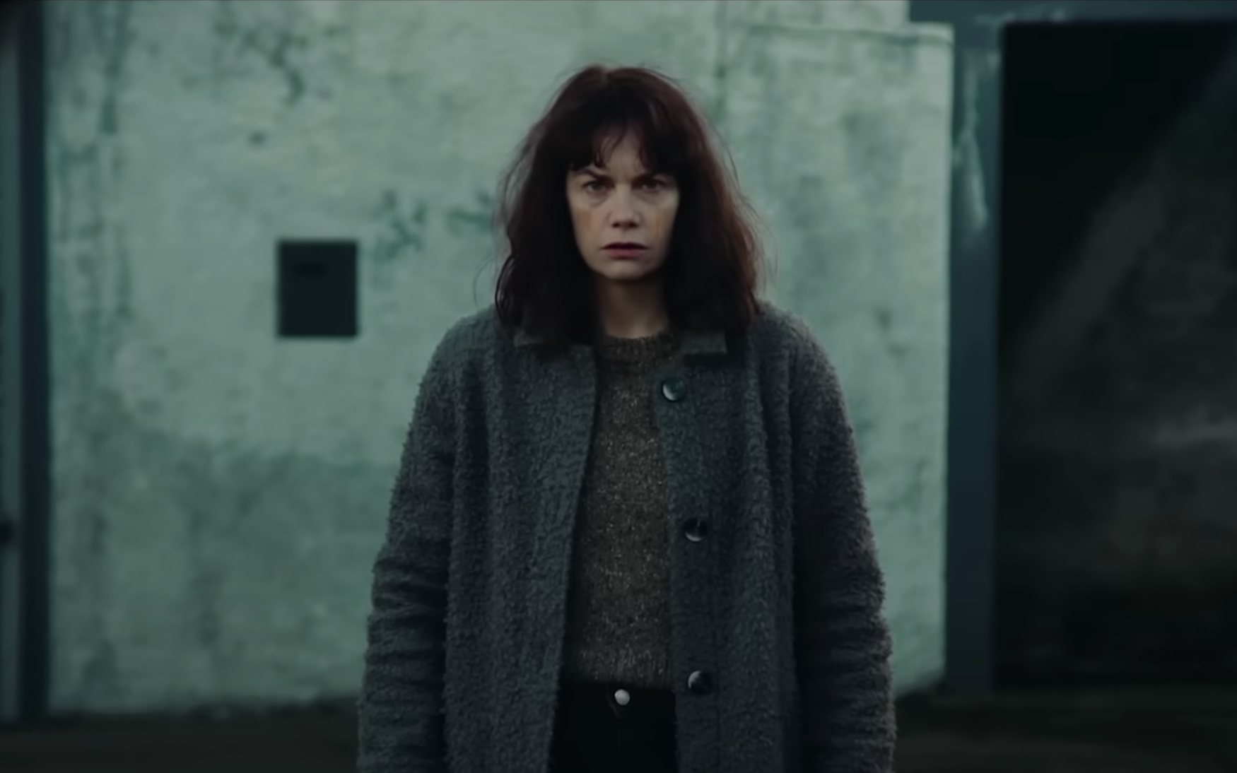 A disheveled woman wearing grey oversized clothes stands in the street staring straight ahead with a confused but resigned expression.