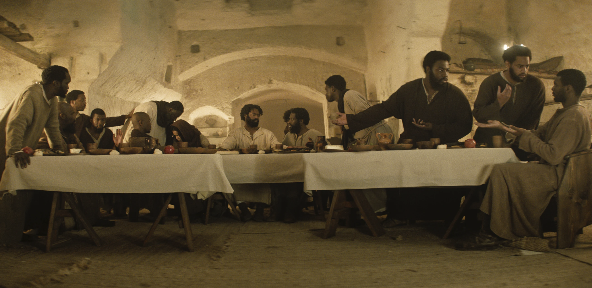 A white stone room with tables arranged. It resembles the Last Supper, but with a Black messiah and disciples. 