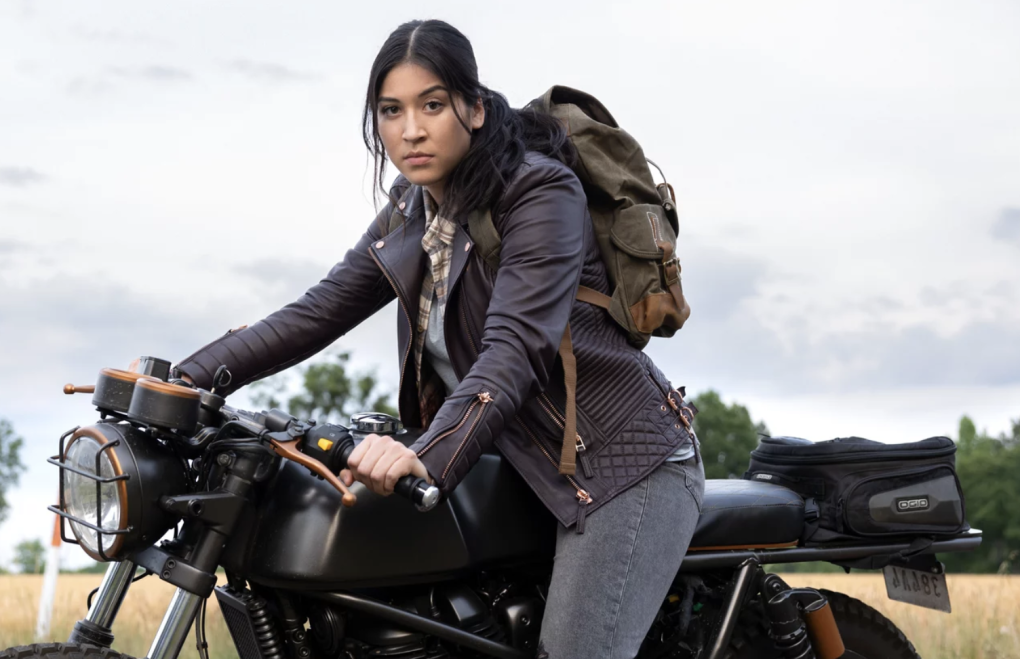 A young woman of color sits astride a black motocycle. She is wearing black jeans, a leather jacket, a backpack and no helmet.