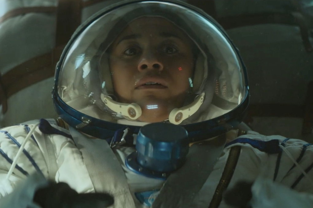 A woman of color wears a nervous expression from inside an astronaut uniform and helmet.