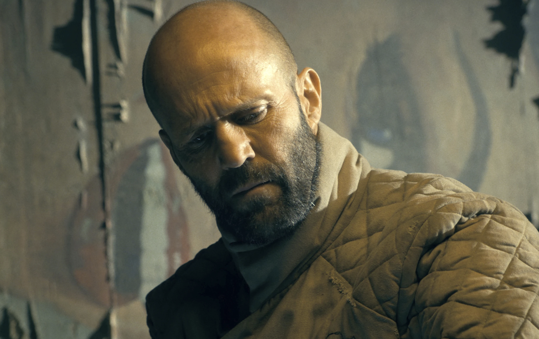 A white bald man in his 50s, with stubble on his face, wearing a padded coat looks down, forehead in a frown.