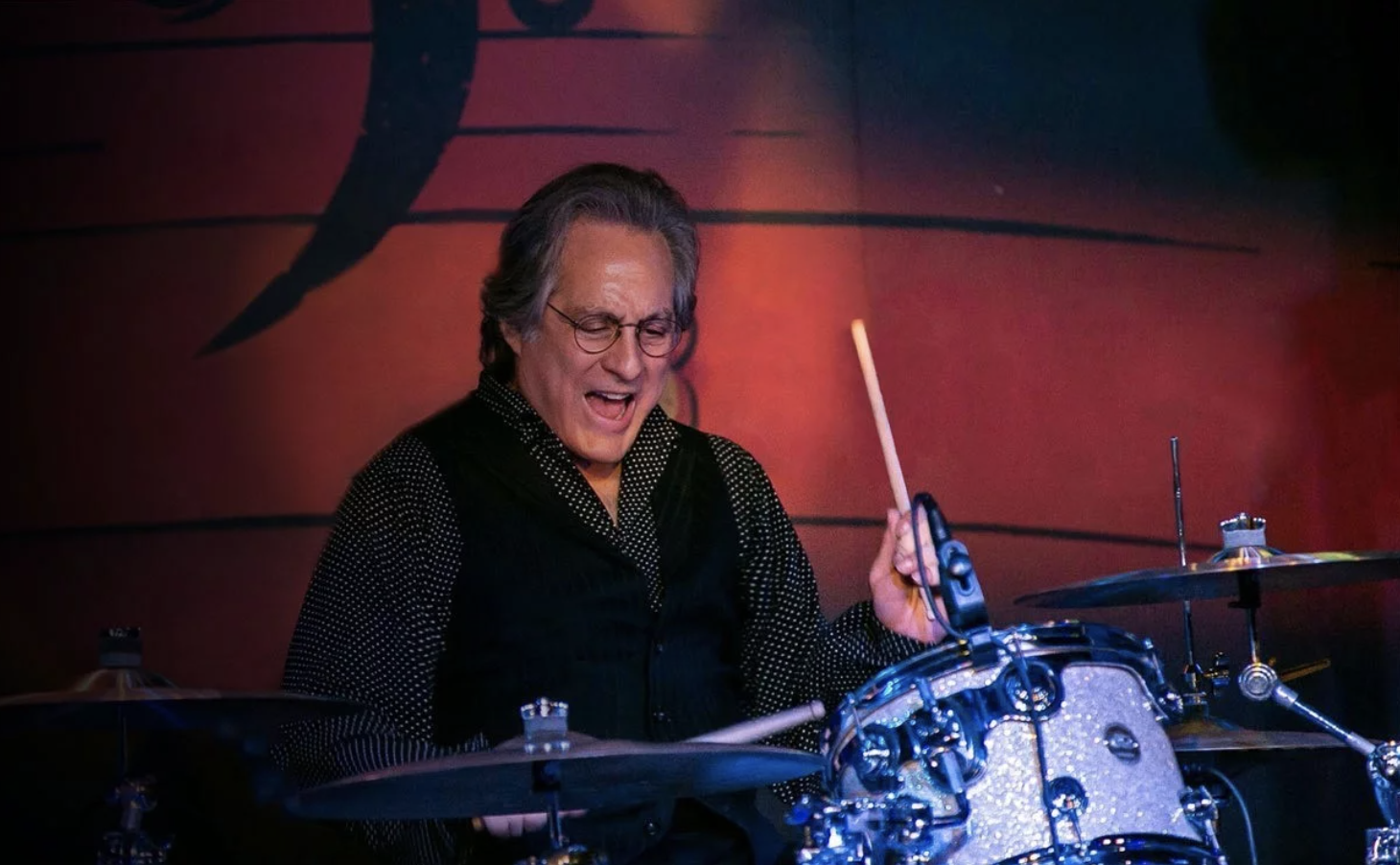 A senior man wearing round spectacles cries out enthusiastically while playing the drums.