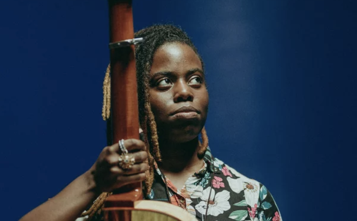 A Black woman wearing a Hawaiian shirt stands clutching the neck of a guitar, while looking off to one side.