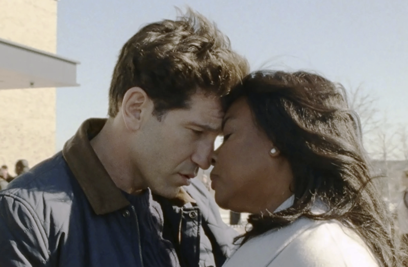 A white man and a Black woman stand outdoors with their foreheads touching, forlornly.