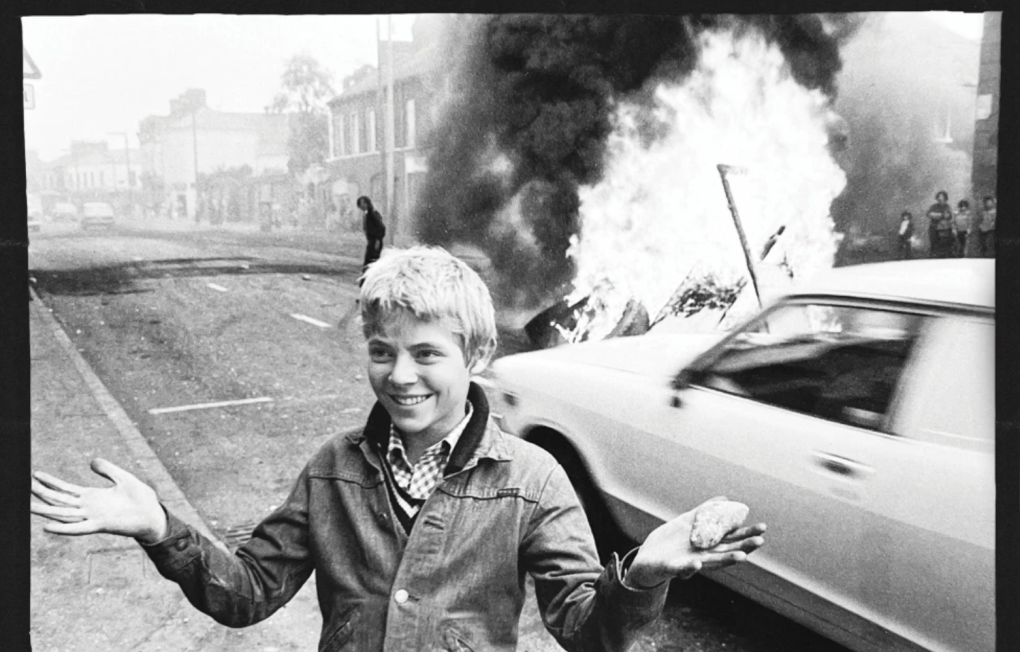 A young person stands smiling next to a car that's on fire and billowing smoke.