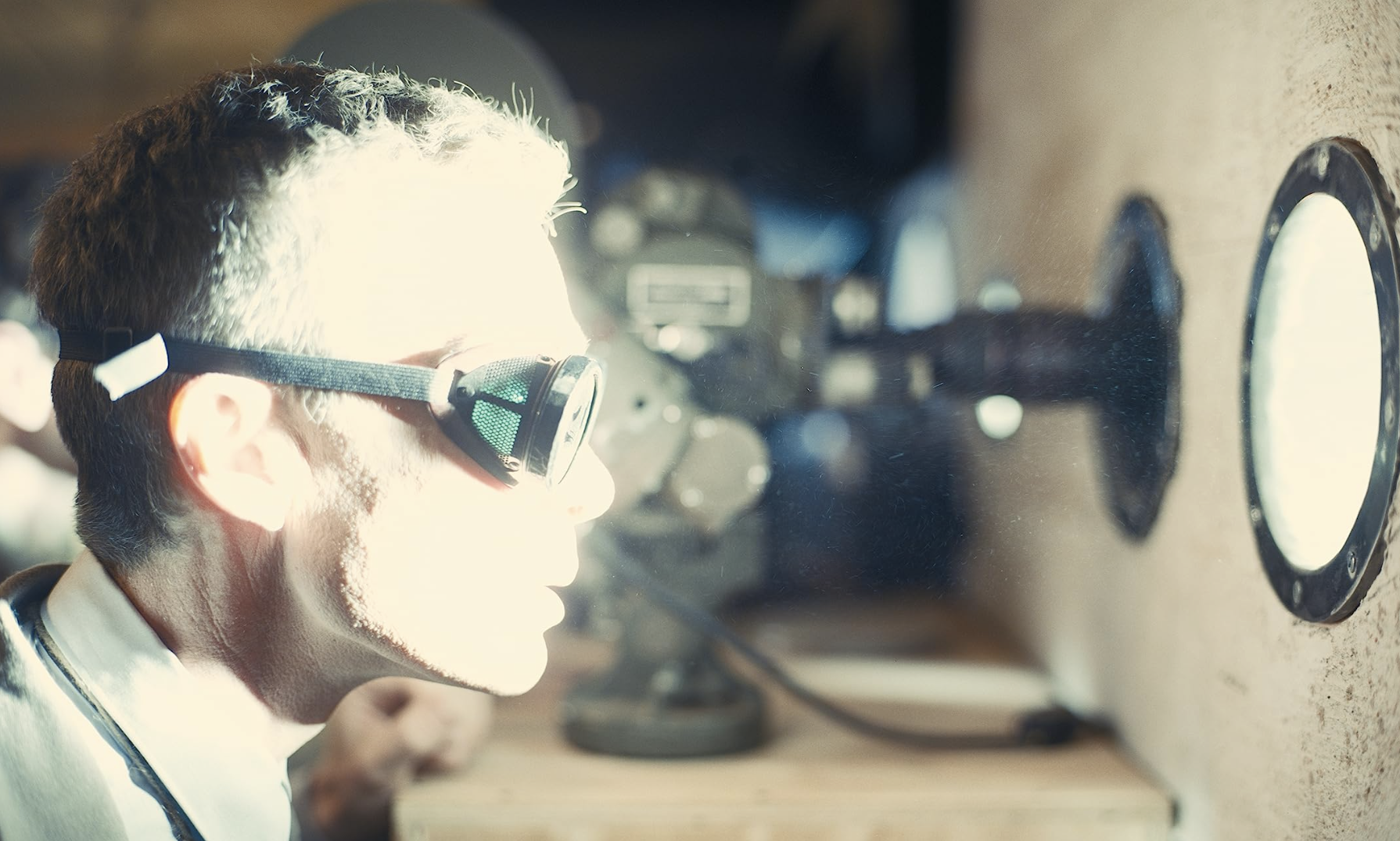 A man viewed in side profile, wearing goggles and staring through a small round window. His face is brightly lit.