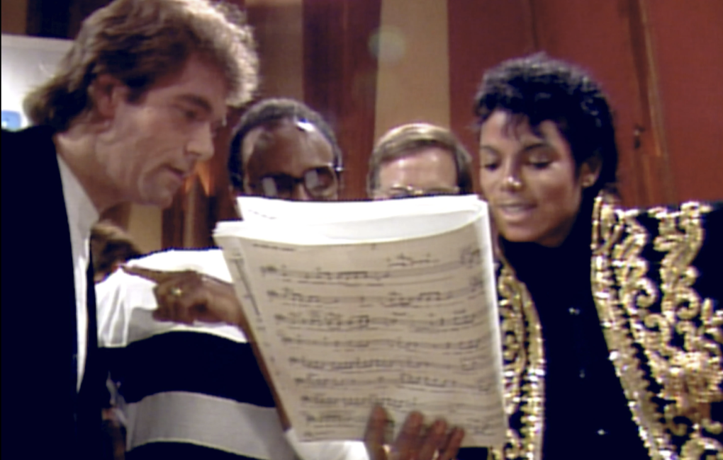 Two white men and two Black men gather around sheet music inside a recording studio.