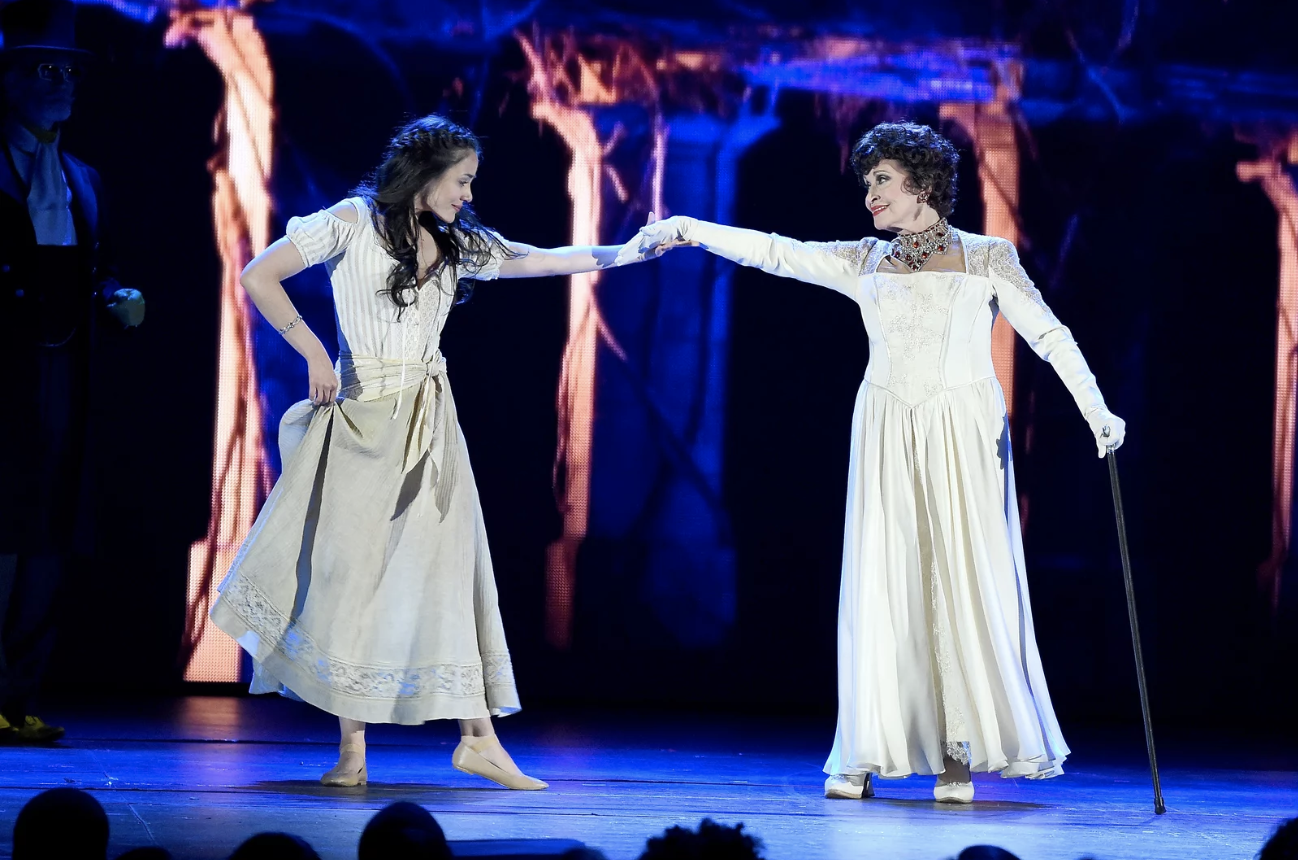 A young woman and an older woman stand on stage, arms outstretched towards one another.