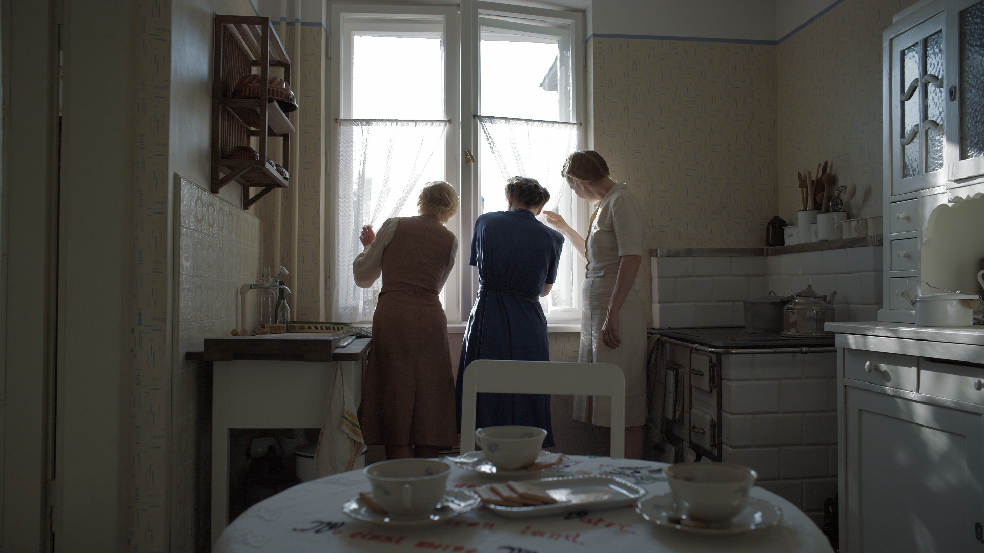 Three women look out through curtains in a kitchen