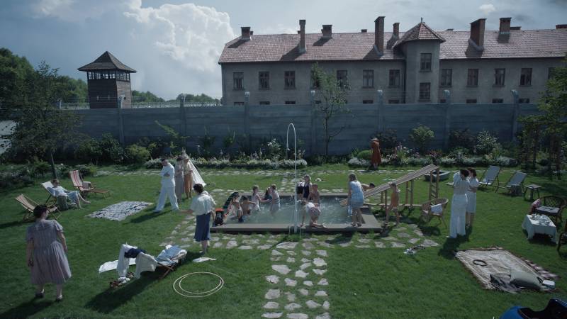 A pool party with children and parents with barbed-wire-topped wall separating grassy yard from institutional building