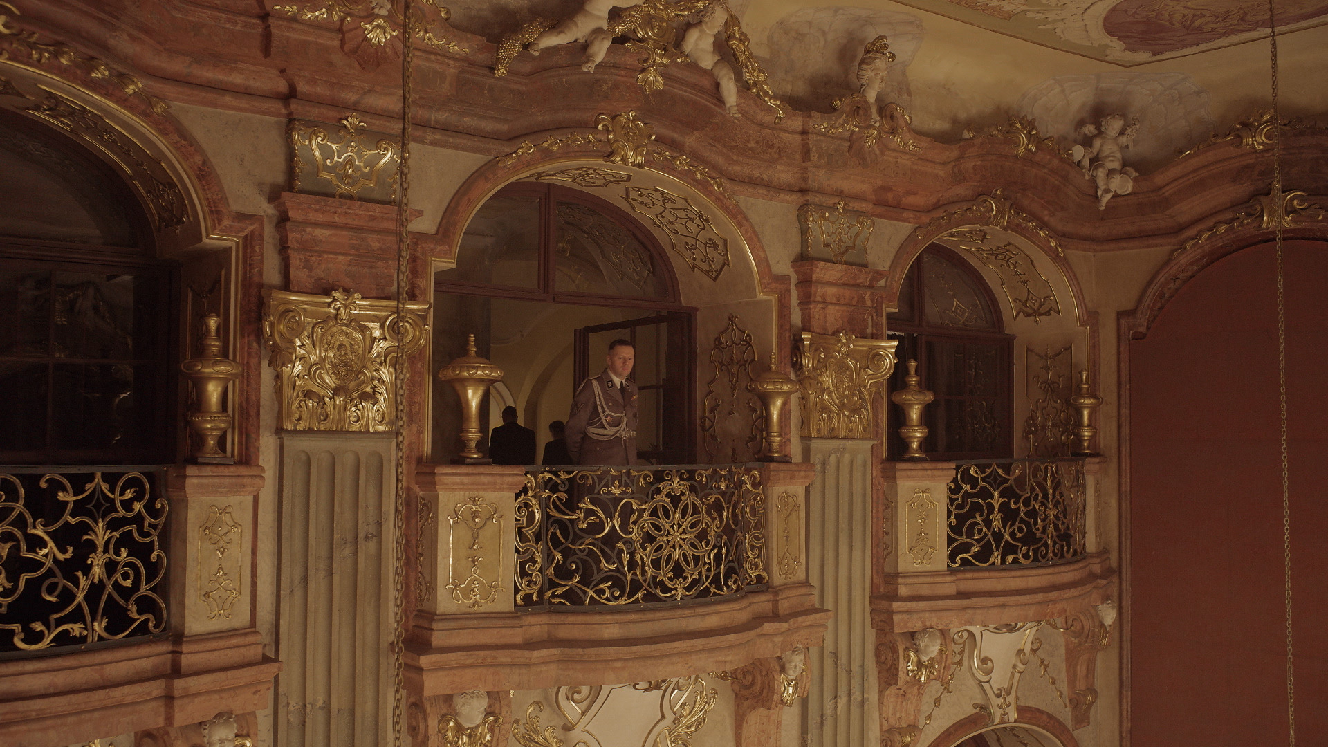 Man in uniform looks out from ornate balcony in gilded, heavily decorative space