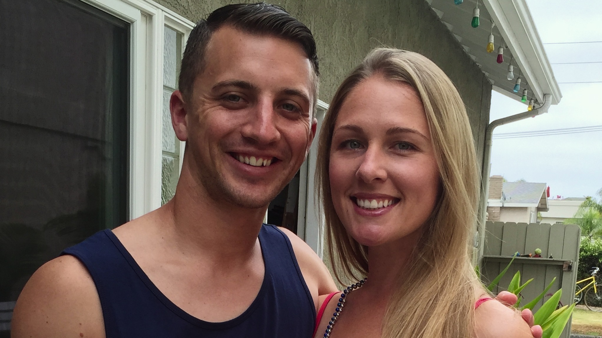 A young attractive man and woman stand close together and smiling outside a home.
