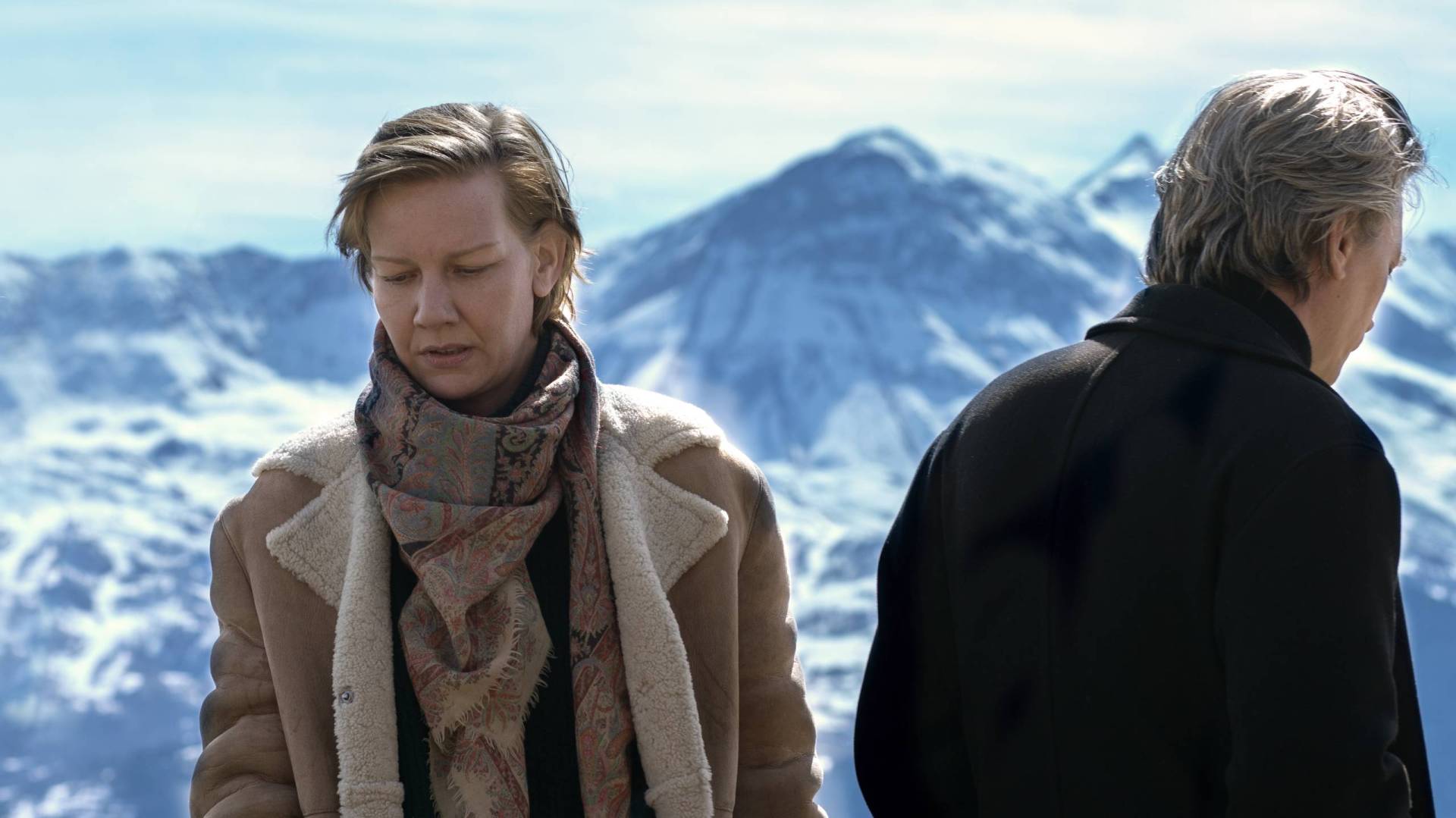 A white woman stands wearing warm clothing, a snow-capped mountain in the distance. A man is at her side, turned away from her.