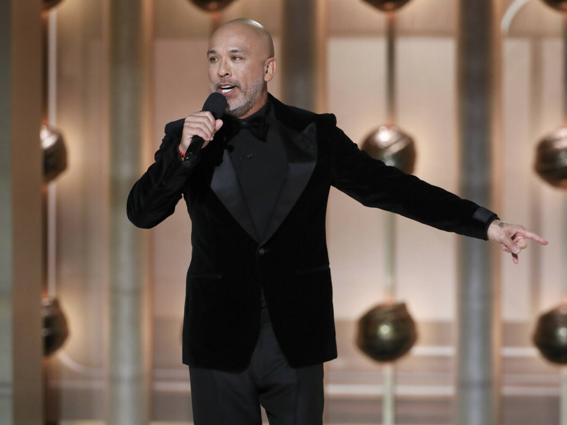 A bald man in a black suit and shirt gestures while speaking into a microphone on stage.