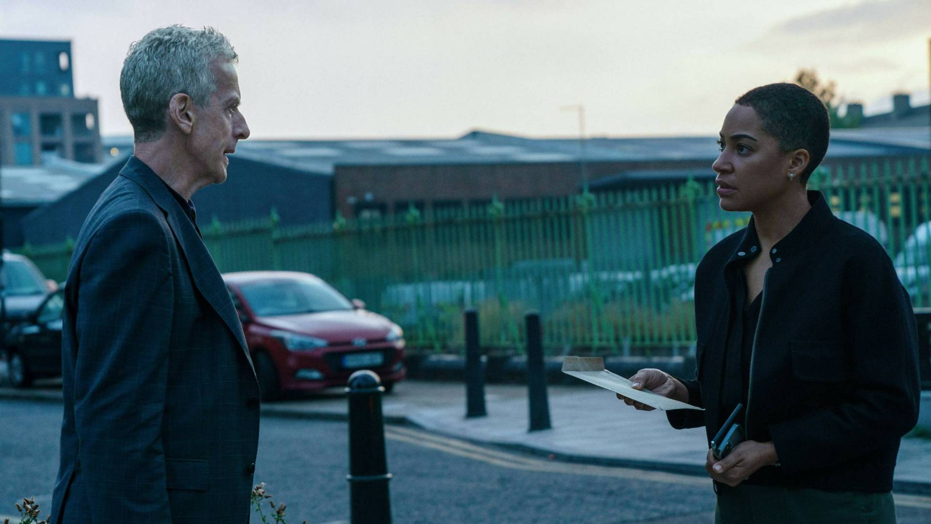 An older white man and younger Black woman talk in the street, near a British industrial estate.