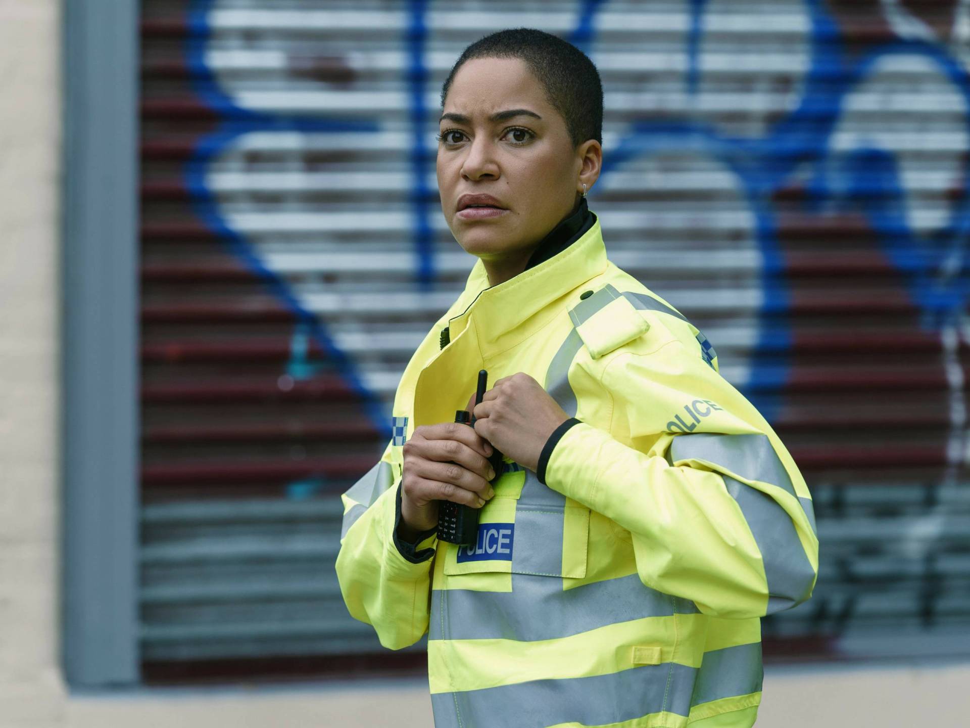 A Black female police officer with a shaved head stands in front of a graffiti'd shop front.