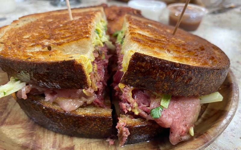 A pastrami sandwich on thick, griddled rye bread, served on a faux wood plate.