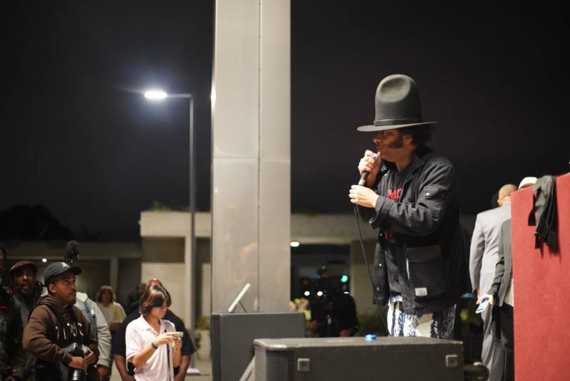Boots Riley wears a signature hat and holds a mic as he stands on stage at the Oakland Museum of California during an event honoring the 50th anniversary of hip-hop.