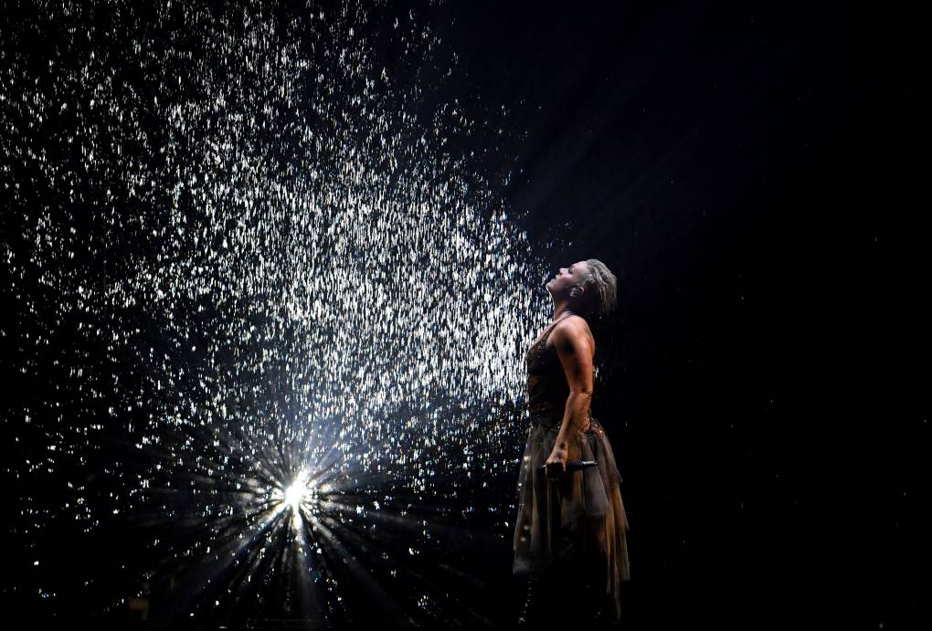 A woman stands on a dark stage getting showered with spot-lit droplets of water.