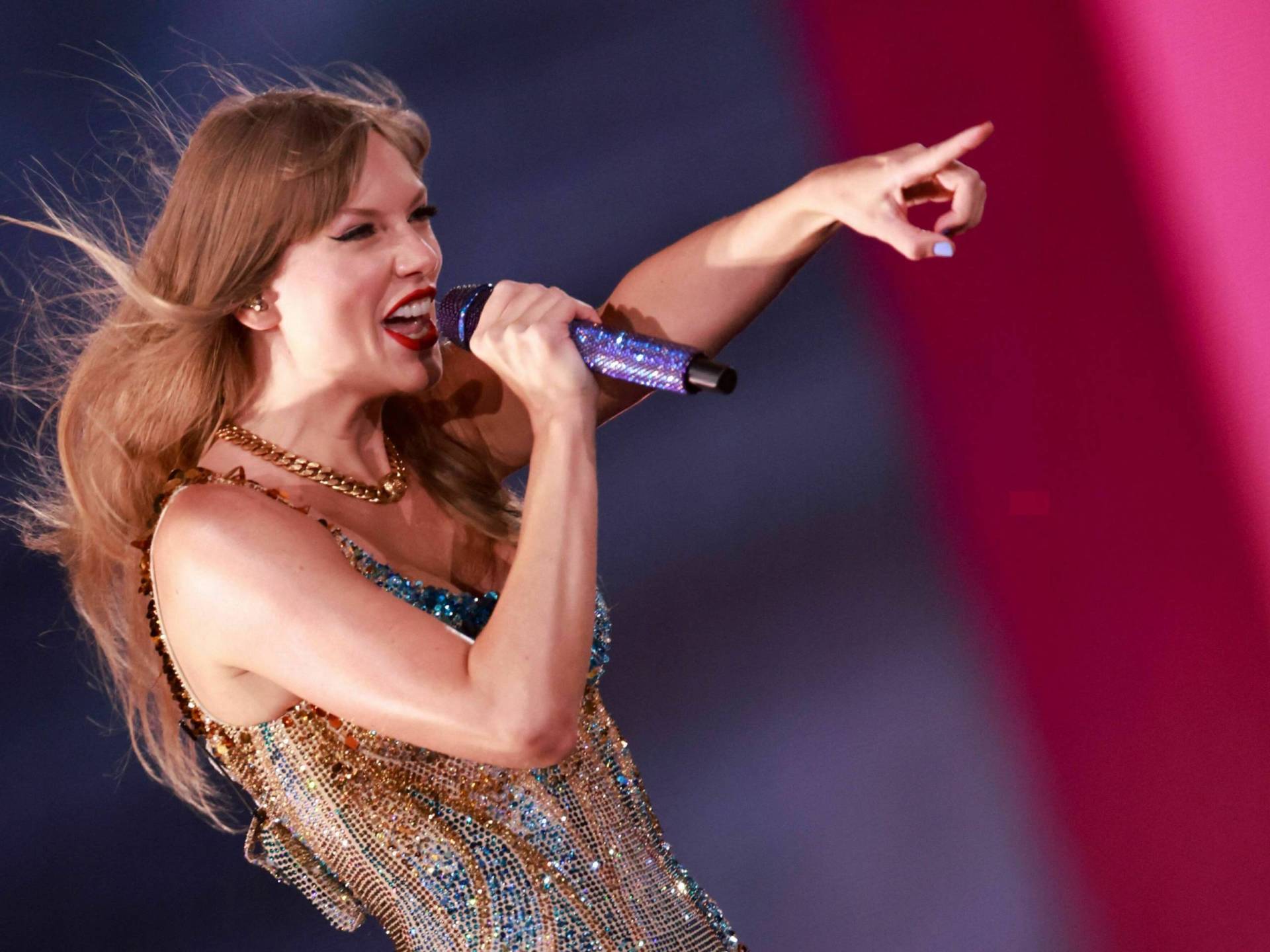 A woman in a sequined body suit stands on stage singing into a microphone and pointing one arm ahead.