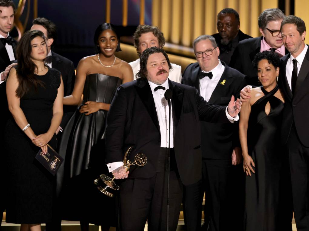 Men and women of various ages and races crowd onto a stage behind a large white man with a mustache who is clutching a trophy.