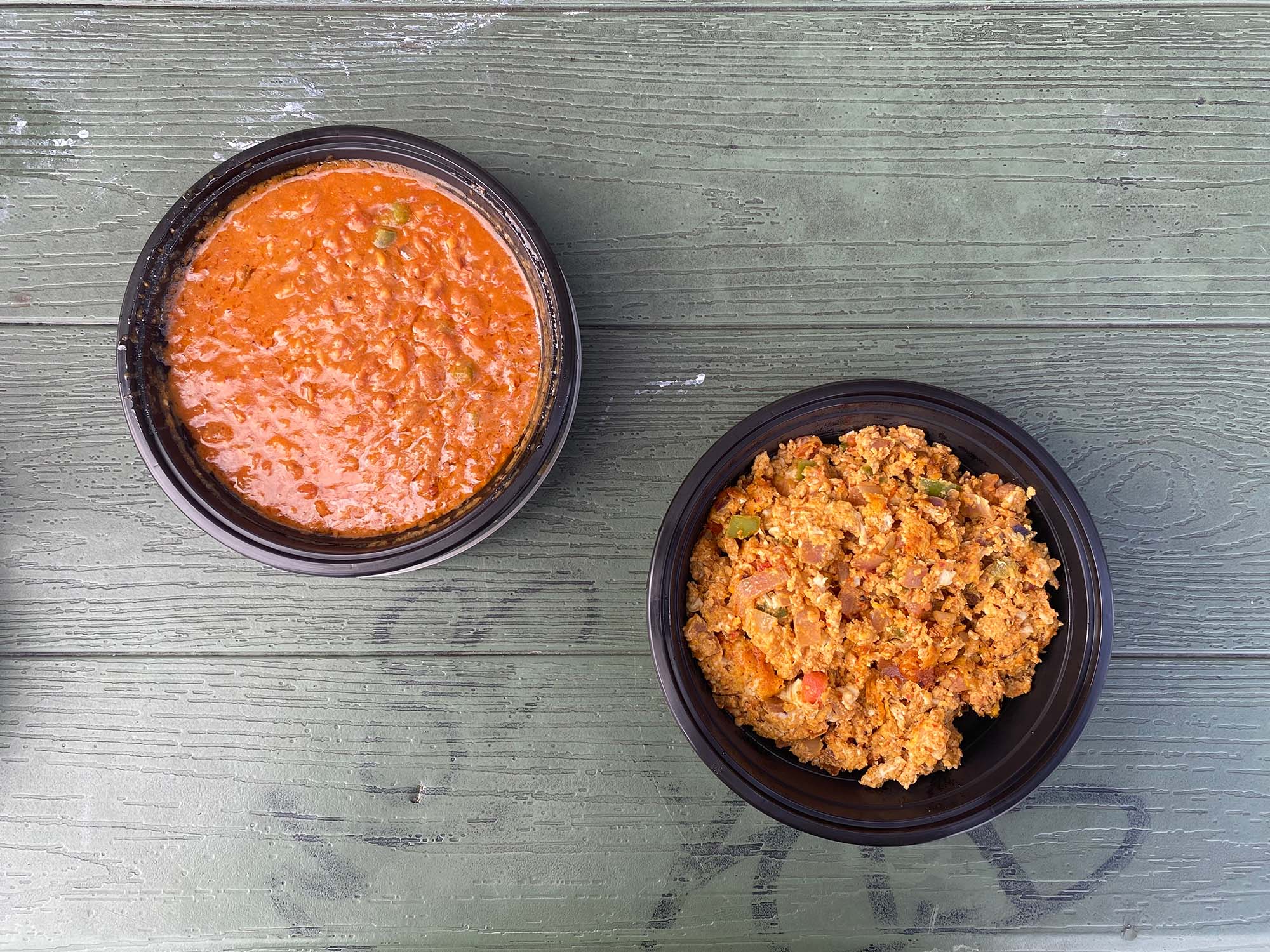 Two round plastic takeout containers on a picnic table. To the left is foul, a soupy, orange-hued bean stew. To the right is the shakshouka eggs, a kind of chunky vegetable scramble.