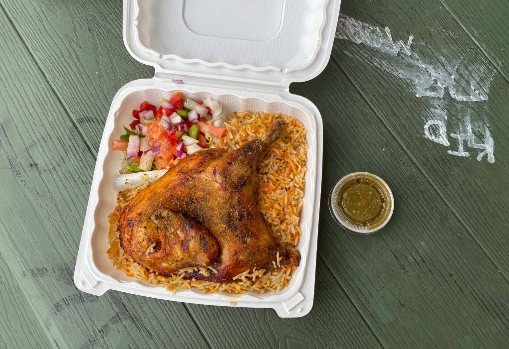On a picnic table: half a roast chicken over a bed of orange-tinted seasoned rice, served in a plastic takeout container with a small tub of green hot sauce on the side.