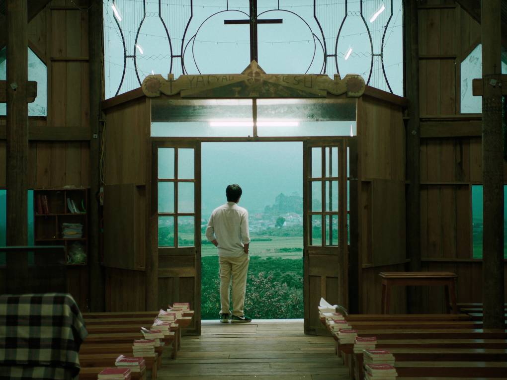 A man stands framed in the doorway of a church entrance. He is gazing out over green and misty fields.