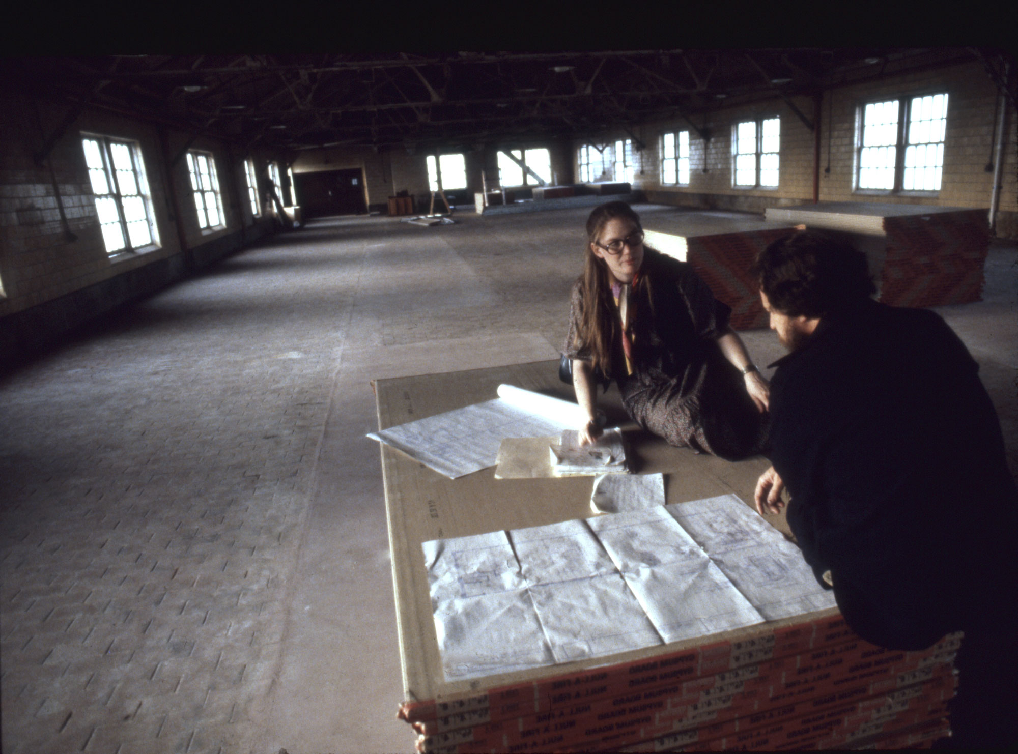 Two people sit on stack of drywall in a cavernous warehouse space looking at architectural plans